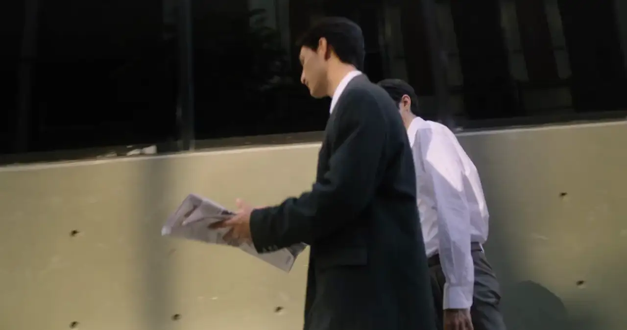 Two men in office clothes walking past a building, one reading a newspaper.